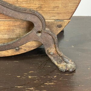 May include: Close-up of an antique wooden barrel with a metal support. The barrel has a curved wooden body and is supported by a decorative, weathered metal leg. The wood has a natural grain.