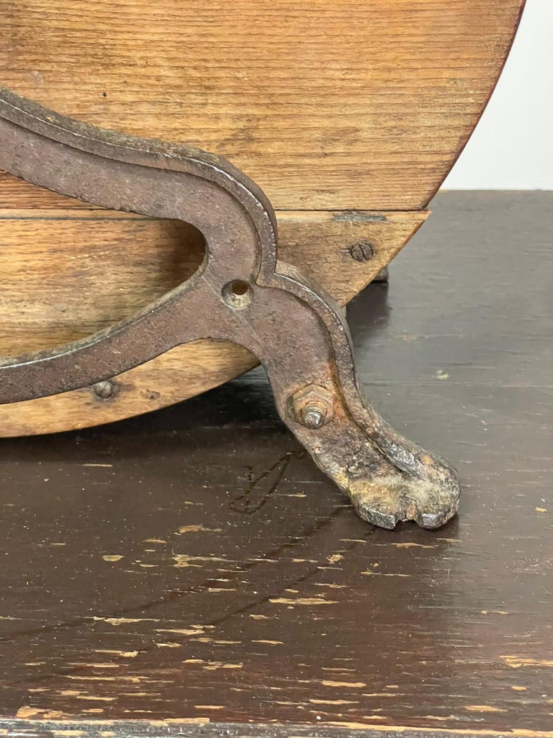 May include: Close-up of an antique wooden barrel with a metal support. The barrel has a curved wooden body and is supported by a decorative, weathered metal leg. The wood has a natural grain.