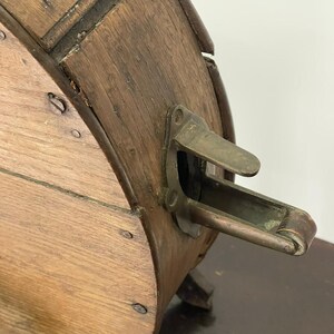 May include: Close-up of an antique wooden barrel with metal hardware. The barrel is made of light brown wood with dark brown accents and metal rivets. A brass-colored metal latch is visible on the side. The barrel is likely used for storage or decoration.