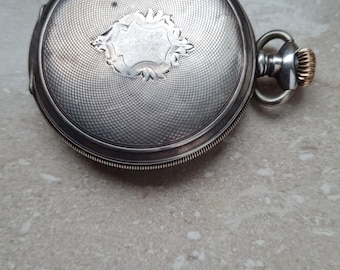 Puede incluir: Reloj de bolsillo de plata con un diseño decorativo en la parte posterior. El reloj tiene una corona de cuerda dorada.