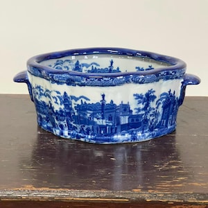 May include: An oval ceramic bowl featuring a blue and white transferware pattern depicting a detailed landscape with buildings, trees, and figures. The bowl has a scalloped rim, two side handles, and a deep basin with a continuous blue design.