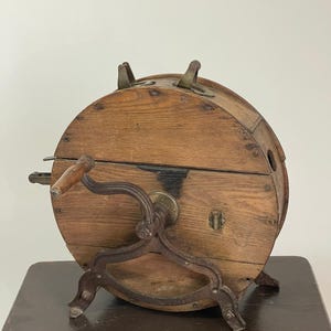 May include: An antique wooden butter churn with a hand crank and metal accents. The churn has a cylindrical shape with a wooden exterior and metal legs. The hand crank is attached to the side, and the churn sits on a dark wooden table.
