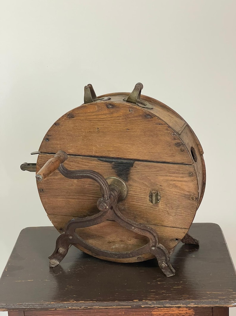 May include: An antique wooden butter churn with a hand crank and metal accents. The churn has a cylindrical shape with a wooden exterior and metal legs. The hand crank is attached to the side, and the churn sits on a dark wooden table.
