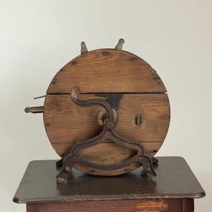 May include: Antique wooden butter churn with a circular design and metal accents. The churn features a hand crank, a metal stand, and a small lock. The wooden body is a warm brown color, and the metal components have a dark, aged appearance.