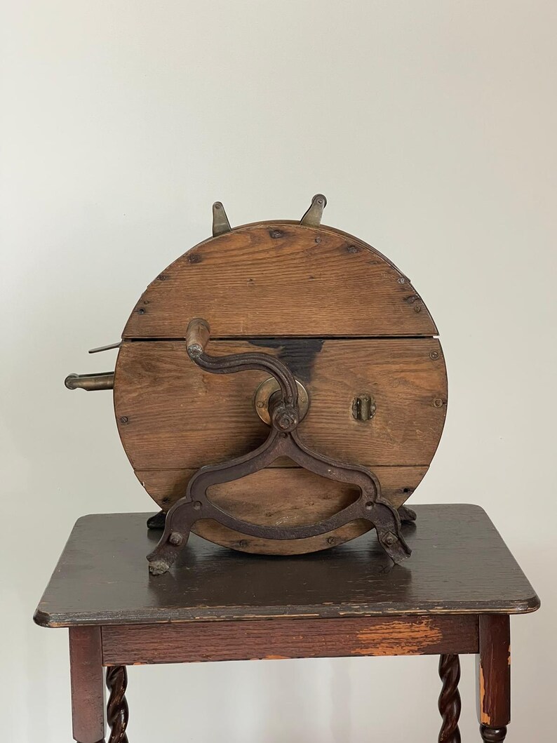 May include: Antique wooden butter churn with a circular design and metal accents. The churn features a hand crank, a metal stand, and a small lock. The wooden body is a warm brown color, and the metal components have a dark, aged appearance.