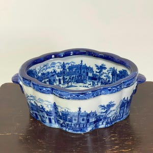 May include: An oval ceramic bowl featuring a blue and white transferware pattern of architectural structures, columns, and figures. The bowl has a scalloped rim, two small side handles, and a continuous landscape scene wrapping around the exterior.