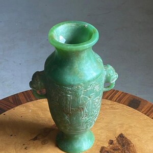 Puede incluir: Un jarrón de jade verde con boca ancha y dos asas. El jarrón presenta detalles en relieve tallados de figuras y arquitectura. Se exhibe sobre una mesa de madera con borde decorativo.