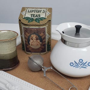 Op de afbeelding: Een witte theepot met een blauw bloemenmotief, een bruine keramische mok, een metalen theezakje en een vintage blikje met Lipton-thee.