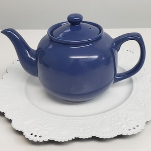 May include: A blue ceramic teapot with a lid, sitting on a white lace doily.