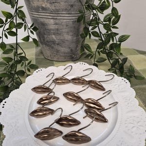 May include: A set of twelve bronze-coloured metal shower curtain hooks, each featuring a three-dimensional lip-shaped design. The hooks are arranged in a circular pattern on a white decorative plate with a scalloped edge and embossed details.