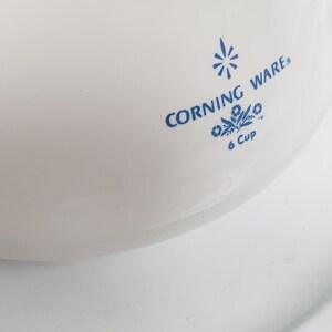 Op de afbeelding: Witte Corning Ware 6-kops ovenschaal met blauwe tekst en een bloemenmotief.