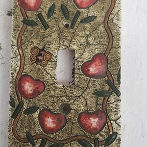 Peut inclure: Un cache-interrupteur décoratif avec un motif de cœurs rouges, de feuilles vertes et d'une vigne brune sur un fond beige. Le cache-interrupteur est doté d'une plaque d'interrupteur blanche.