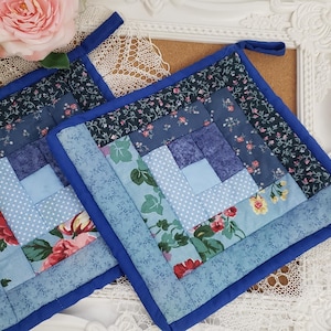 Puede incluir: Dos salvamanteles cuadrados acolchados con un diseño floral y patchwork azul. Los salvamanteles tienen un borde azul y un lazo para colgar. El patchwork incluye telas florales, de lunares y azul liso.