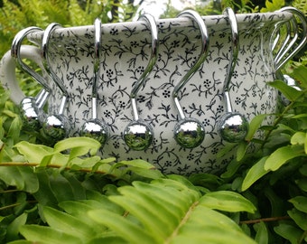 May include: A set of 12 silver shower curtain hooks with a round ball at the end of each hook. The hooks are displayed in a white ceramic bowl with a black floral pattern.