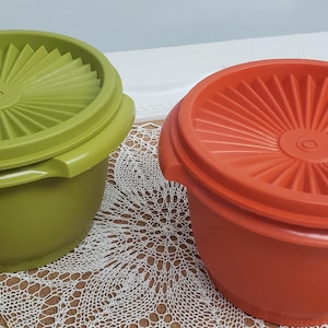 Puede incluir: Dos cuencos Tupperware vintage, uno verde oliva y otro naranja, cada uno con una tapa a juego. Las tapas tienen un diseño radial. Los cuencos están sobre un mantelito decorativo.