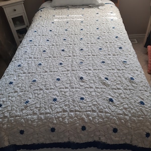 Peut inclure: Une courtepointe blanche en crochet avec des fleurs brodées bleues et une bordure en crochet bleue. La courtepointe est sur un lit simple avec un oreiller blanc.
