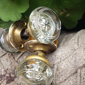 Peut inclure: Plusieurs boutons de porte anciens en verre avec des bases en laiton. Les boutons en verre transparent présentent un motif en forme d'étoile. Les boutons de porte sont posés sur une surface en pierre grise texturée, avec des feuilles vertes en arrière-plan.