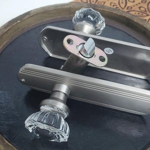 Puede incluir: Dos manillas de puerta plateadas con pomos de cristal transparente. Las manillas están montadas sobre una superficie de madera con un patrón decorativo.