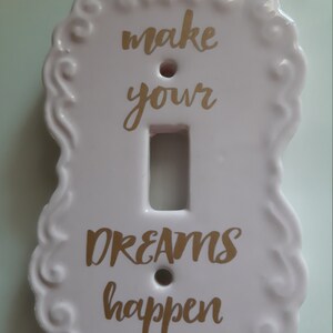 以下が含まれることがあります： 「make your dreams happen」という金色の文字が書かれた白いセラミック製のライトスイッチカバーです。