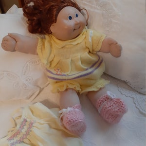 Könnte beinhalten: Eine Cabbage Patch Kid Puppe mit roten Locken, die ein gelbes Strickkleid mit lila Besatz und rosa gehäkelte Stiefelchen trägt. Die Puppe liegt auf einem weißen Bettlaken mit Blumenmuster.