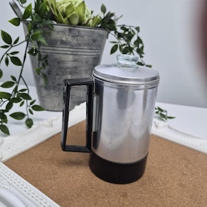 May include: A vintage aluminum coffee percolator with a black base and handle. The percolator has a glass top and is cylindrical in shape. The image also shows a galvanized metal bucket with greenery.