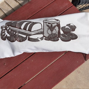 May include: White cotton drawstring bag with a brown print of a loaf of bread, a bag of flour, and some rolls. The bag is lying on a red wooden surface.