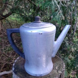 May include: A silver aluminium coffee pot with a black handle and a black lid. The pot is sitting on a tree stump.