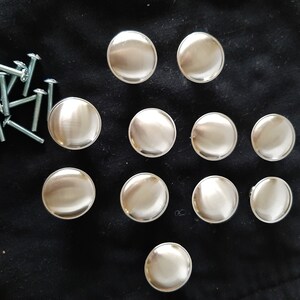 K&ouml;nnte beinhalten: Zw&ouml;lf runde, silberfarbene Schrankkn&ouml;pfe mit glatter, gl&auml;nzender Oberfl&auml;che. Die Kn&ouml;pfe sind in einem Kreis auf einer schwarzen Oberfl&auml;che angeordnet.