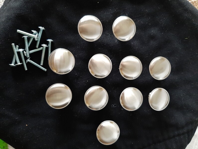 K&ouml;nnte beinhalten: Zw&ouml;lf runde, silberfarbene Schrankkn&ouml;pfe mit glatter, gl&auml;nzender Oberfl&auml;che. Die Kn&ouml;pfe sind in einem Kreis auf einer schwarzen Oberfl&auml;che angeordnet.