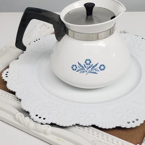Op de afbeelding: Witte theepot van keramiek met een blauw bloemenmotief en een zwarte handgreep. De theepot staat op een wit kantonderzetter.