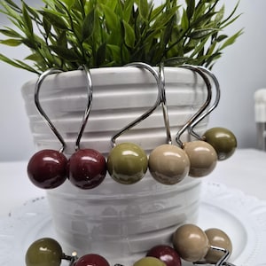 May include: A set of shower curtain hooks with decorative, round, colored accents. The hooks are silver-toned metal with a curved design. The accents are in shades of burgundy, olive green, and beige. The hooks are displayed near a white ceramic pot with greenery.