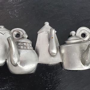 May include: A collection of five silver-colored teapots, each with a unique design, arranged in a row. The teapots vary in shape, with spouts, handles, and lids. The background is a dark, textured surface.