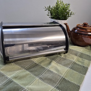 May include: A stainless steel bread bin with a roll-top lid, beside a brown ceramic teapot and a small potted plant. The bread bin is rectangular with rounded edges and a black trim. The teapot has a lid and a handle.