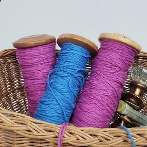 May include: A wicker basket contains three yarn spools: two magenta and one blue. The spools have wooden tops. Vintage glass doorknobs and keys are also in the basket. The basket has a woven handle.
