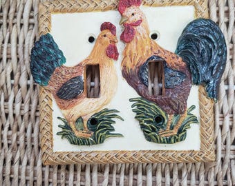 Può includere: Una placca per interruttore della luce decorativa con due galli, uno marrone e uno bianco e nero, con erba verde e un bordo intrecciato marrone.