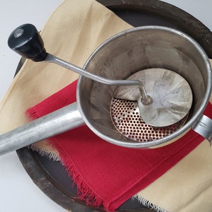 May include: A vintage silver food mill with a black handle. The mill has a round bowl with a perforated base and a rotating blade.