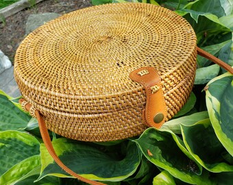 May include: A round, woven straw bag with a brown leather strap and a brass buckle. The bag is sitting on a bed of green leaves.