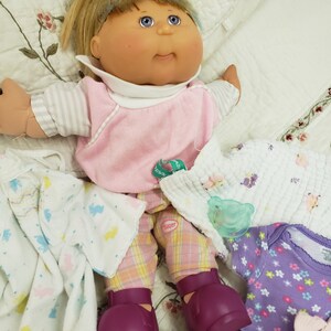 Op de afbeelding: Een Cabbage Patch Kid pop die een roze en wit gestreept shirt, een roze broek met een ruitpatroon en paarse schoenen draagt. De pop is omringd door verschillende babykleding en accessoires, waaronder een wit rompertje met een bloemenpatroon, een wit deken met konijnen, een paars rompertje met een bloemenpatroon en een groen en wit deken.