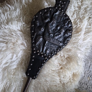 May include: A black fireplace bellows featuring a relief-moulded scene of figures gathered around a table. The bellows has a dark, smooth surface secured with numerous metal studs along the edges and a long, narrow metal nozzle at the bottom end.