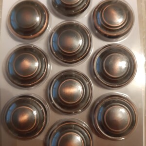 Może przedstawiać: Dziesięć okrągłych, brązowych gałek do szafek z wypukłym wzorem koncentrycznych k&oacute;ł. Gałki są zapakowane w przezroczystą folię plastikową z nadrukowaną nazwą marki "Amerock" i frazą "&iexcl;VALOR ECONOMICO!"