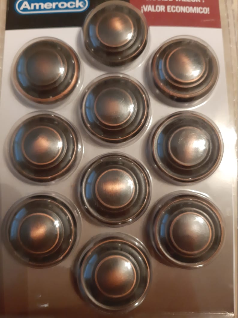 Może przedstawiać: Dziesięć okrągłych, brązowych gałek do szafek z wypukłym wzorem koncentrycznych k&oacute;ł. Gałki są zapakowane w przezroczystą folię plastikową z nadrukowaną nazwą marki "Amerock" i frazą "&iexcl;VALOR ECONOMICO!"