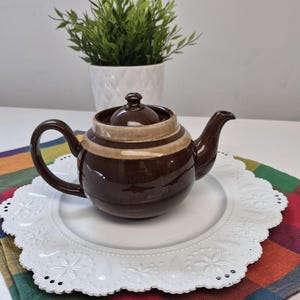 Peut inclure: Une théière en céramique brun foncé brillant avec un corps arrondi, une anse incurvée et un couvercle assorti. La théière présente une large bande horizontale beige clair sur la partie supérieure et repose sur une assiette décorative blanche.
