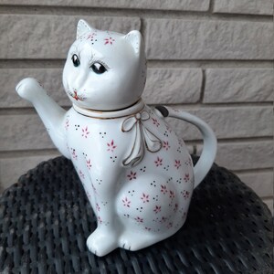 May include: White ceramic teapot shaped like a cat with pink and black floral pattern. The teapot has a gold ribbon around the neck and a white lid with a cat face.