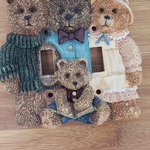 Könnte beinhalten: Eine Lichtschalterabdeckung mit vier braunen Teddybären. Die Bären tragen verschiedene Outfits, darunter einen grünen Pullover, ein blaues Hemd mit einer braunen Fliege, ein weißes Kleid mit rosa Punkten und eine blaue Latzhose. Die Bären sind in einer Reihe angeordnet, wobei die beiden größeren Bären außen und die beiden kleineren Bären in der Mitte sind.