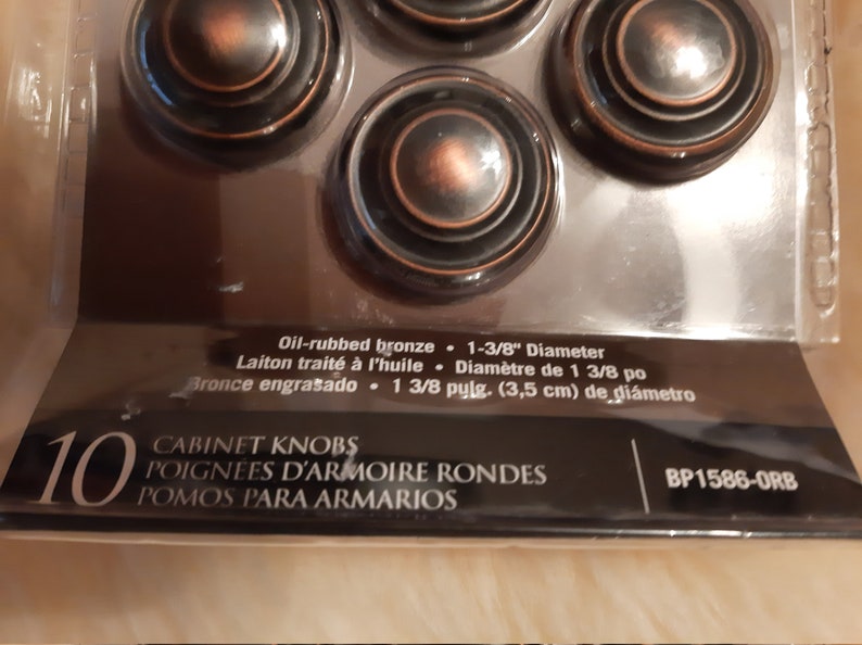 Może przedstawiać: Opakowanie 10 gałek meblowych z brązu olejowanego. Gałki są okrągłe i mają średnicę 3,5 cm. Opakowanie zawiera r&oacute;wnież tekst "CABINET KNOBS, POIGNEES D'ARMOIRE RONDES, POMOS PARA ARMARIOS, BP1586-0RB".