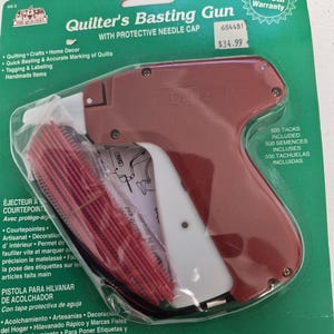 May include: A Dritz Quilter's Basting Gun with a protective needle cap, packaged on a green card. The tool is maroon and white, featuring a trigger mechanism. Included are 500 red plastic tacks. The packaging displays the product name and text.