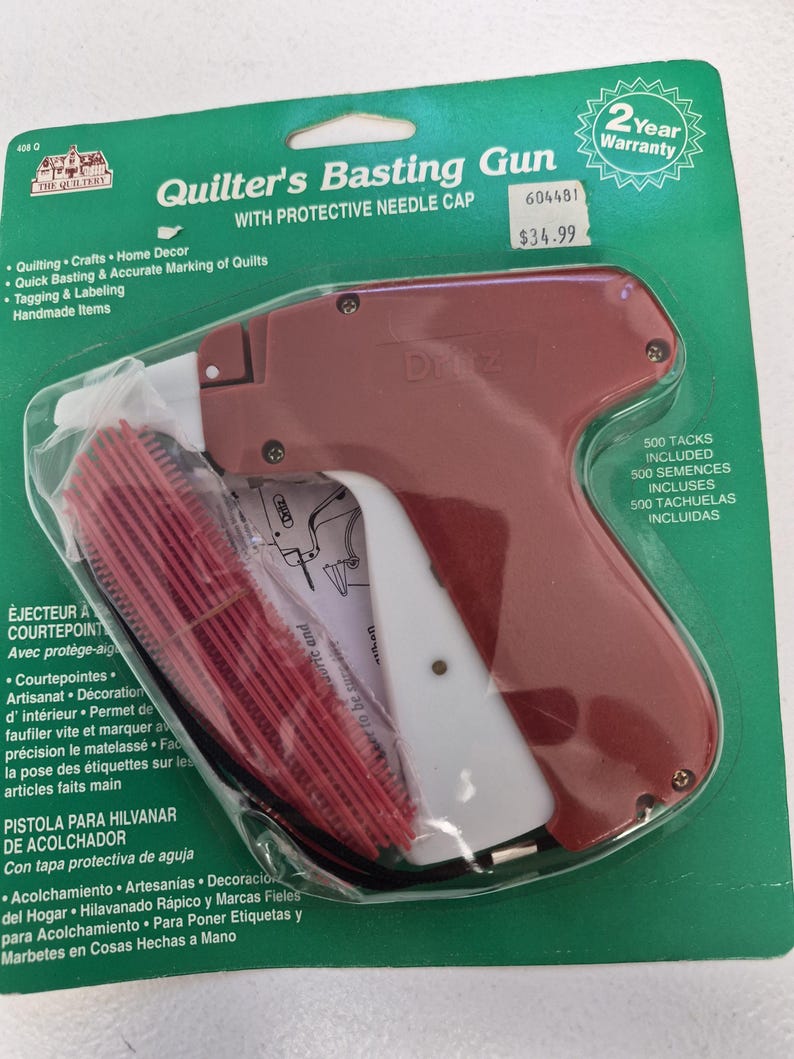 May include: A Dritz Quilter's Basting Gun with a protective needle cap, packaged on a green card. The tool is maroon and white, featuring a trigger mechanism. Included are 500 red plastic tacks. The packaging displays the product name and text.