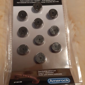 Może przedstawiać: Opakowanie dziesięciu czarnych gałek do szafek z dopasowaną gałką do szafki. Opakowanie zawiera 10 śrub do łatwego montażu. Opakowanie jest oznaczone "Amerock" i "BP1584-ORB".