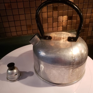 May include: A silver aluminum kettle with a black handle and a black lid. The kettle has a small spout and a label that reads "FOR USE WITH GAS OR ELECTRIC STOVES ONLY.  DO NOT USE ON DIRECT HEAT.  REMOVE FROM HEAT BEFORE POURING.  HANDLE WITH CARE.  MADE IN JAPAN."