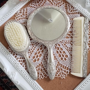May include: A white vanity set with a silver hand mirror, a silver hairbrush, and a white comb. The set is displayed on a cork board with a white frame and a lace doily.